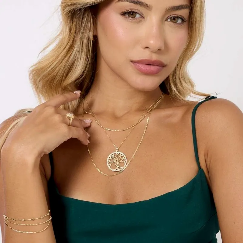 Modelo de cabelo loiro e vestido verde, com três colares dourados, um deles com pingente de árvore da vida, anel e pulseira dourados
